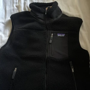 Svart fleeceväst från Patagonia M - Svart fleeceväst från Patagonia i storlek M. Västen har hög krage, tre fickor med dragkedja och en bröstficka med logotyp. Tillverkad i mjuk fleece och detaljer i syntetmaterial. Perfekt för lager-på-lager och outdoor.