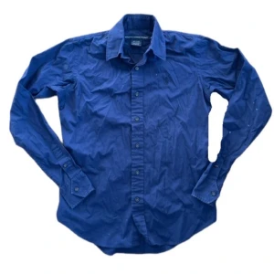 Blå skjorta från Ralph Lauren Sport - Klassisk blå skjorta från Ralph Lauren Sport med broderad logga på bröstet. Skjortan har lång ärm, knappar framtill och är tillverkad i mjuk bomull med lite stretch för extra komfort. Perfekt för dig som gillar stilren och tidlös design.