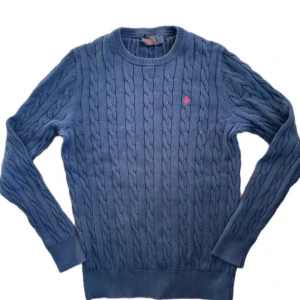 Blå kabelstickad tröja från Morris - Kabelstickad blå tröja från Morris med rund hals och diskret röd broderad logga på bröstet. Tröjan är i mjuk bomull, har långa ärmar och ribbade muddar. Perfekt för dig som gillar klassisk och stilren look.