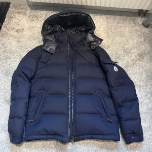 Moncler Montgenevre  - Snygg mörkblå dunjacka från Moncler med avtagbar huva och klassisk logga på ärmen. Små defekter under båda armarna knappt synligt