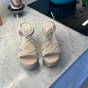 Beiga flätade platåsandaler med kilklack - beiga sandaler från med flätade remmar och hög kilklack.  Skorna har öppen tå, spänne vid ankeln och en plattformssula som ger extra höjd. Perfekta för sommarens alla outfits!