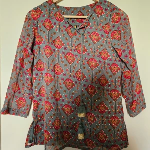 Blommig blus med knytband och tofsar - Säljer en färgglad blus med blommigt mönster i rosa, rött och grönt på grå botten. Blusen har trekvartsärm och dekorativa knytband med tofsar framtill. Perfekt för dig som gillar bohemisk stil och vill sticka ut med coola detaljer.