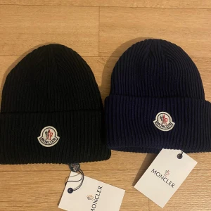Moncler ribbstickade mössor svart & marinblå - Två ribbstickade mössor från Moncler, en i svart och en i marinblå. Båda har uppvikt kant och Monclers klassiska logotyp framtill. Perfekt accessoar för kalla dagar och snygg till streetwear. Materialet är mjukt och värmande, troligen ull eller blandmaterial. Båda för 350kr.