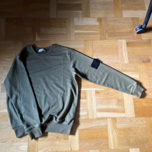 Stone Island olivgrön sweatshirt - olivgrön sweatshirt frän Stone Island med rund hals. Tröjan har den Stone Island-patchen pả vänster ärm, bäde den svarta och den gula patchen kommer med! Har knappt använt den da jag har bytt stil. OBS: Pris kan diskuteras