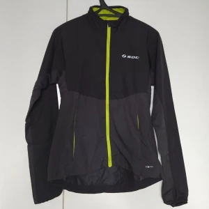 Svart vindjacka från SOC med neon-detaljer - Svart vindjacka från SOC med neon-gula detaljer och dragkedja. Jackan har hög krage, långa ärmar och reflekterande detaljer. Perfekt för löpning eller utomhusaktiviteter. Lätt och smidig modell i syntetmaterial. Vet tyvärr inte storlek då det ej står på jackan, men funkar perfekt för storlek- S & M💗