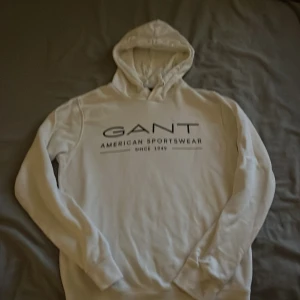 Vit hoodie från GANT med tryck - Vit hoodie från GANT med svart tryck på bröstet där det står 'GANT American Sportswear'. Klassisk modell med huva och långa ärmar. Mjuk insida och ribbade muddar. Perfekt för en avslappnad stil.