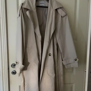 Beige trenchcoat från Bershka - Stilren beige trenchcoat från Bershka med klassisk krage, knappar framtill och snygga detaljer vid ärmsluten. Lång modell med avslappnad passform och stora fickor. Perfekt för dig som vill ha en tidlös och trendig jacka. Pris diskuteras vid snabbaffär!