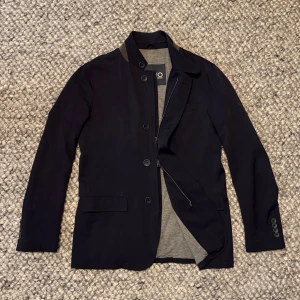 Herno jacka - Stilren mörkblå kavajjacka från Herno med klassisk krage och knäppning framtill. Jackan har grått innerfoder och smart dragkedja under knapparna. Tillverkad i ett tekniskt material med Loro piana Rain System, perfekt för dig som vill ha en dressad look med modern twist.