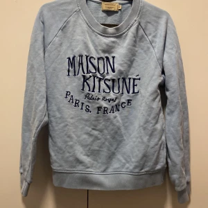 Ljusblå sweatshirt från Maison Kitsuné - Säljer en snygg ljusblå sweatshirt från Maison Kitsuné med tryck på framsidan. Tröjan är i bra skick och perfekt för både vår och höst. Den har långa ärmar och en normal passform. Superbekväm och stilren för alla tillfällen! 