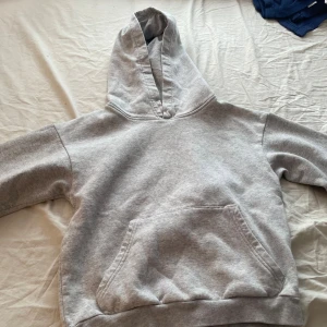 Grå hoodie från H&M med huva - En basic grå hoodie från H&M med stor känguruficka och huva. Tröjan är i mjuk bomullsblandning och har ribbade muddar vid ärmslut och nederkant. Perfekt för chill dagar eller när du vill ha något enkelt och bekvämt den har en liten fläck den går nu säkert ut när man tvättar tröjan.