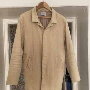 Beige rock i bomull - Stilren beige rock med klassisk krage och raka linjer. Jackan har två snedställda fickor framtill och stängs med dolda tryckknappar. Tillverkad i mjuk bomull, perfekt för övergångssäsonger. Enkel och clean look som passar till det mesta. Sitter riktigt snyggt!