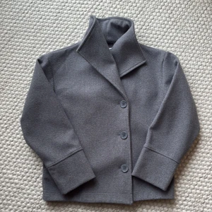 Grå kavajjacka från Gina Tricot 146-152 - Snygg grå kavajjacka från Gina Tricot med klassisk krage och tre knappar framtill. Jackan har lång ärm och är tillverkad i mjukt material som ger en stilren look. Perfekt för dig som gillar minimalistisk och clean stil.