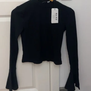 Svart långärmad topp från NA-KD - Snygg svart långärmad topp från NA-KD med utsvängda ärmslut och hög hals. Toppen är croppad och har en slim passform som framhäver figuren. Perfekt att styla med jeans eller kjol för en trendig look.