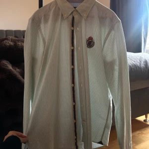 Randig skjorta från Morris, M - Snygg randig skjorta från Morris i ljusgrönt och vitt. Klassisk krage, broderad logga på bröstet och knappar framtill. Skjortan är långärmad och tillverkad i mjuk bomull, perfekt för en stilren look.