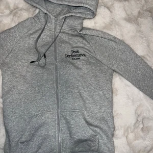 Grå hoodie från Peak Performance - Snygg grå hoodie från Peak Performance med dragkedja framtill och broderad logga på bröstet. Tröjan har huva med snörning och två fickor framtill. Perfekt för en chill och sportig stil.