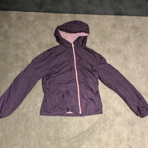 Mörkblå vindjacka från Moncler - Snygg mörklila vindjacka från Moncler med huva och hel dragkedja i kontrastfärg. Jackan har två fickor framtill med knappar och ett klassiskt Moncler-märke på ärmen. Insidan är ljusgrå och materialet är lätt och smidigt, perfekt för blåsiga dagar.