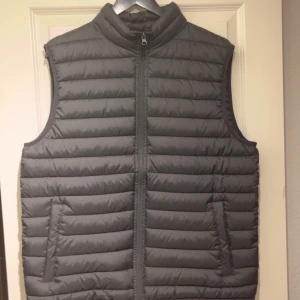 Grå dunväst från Urban Classics XL - Snygg grå dunväst från Urban Classics i storlek XL. Västen har quiltad design, hög krage och dragkedja framtill. Perfekt för lager-på-lager och enkel att matcha med hoodies eller tröjor. Två fickor på sidorna för extra funktionalitet.