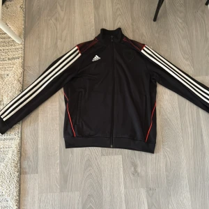 Svart Adidas track jacket med röda detaljer - Svart Adidas track jacket med klassiska vita ränder längs ärmarna, röda detaljer vid axlar och sidor samt hel dragkedja framtill. Jackan har ståkrage, fickor med dragkedja och broderad Adidas-logga på bröstet. Perfekt för en sportig look.