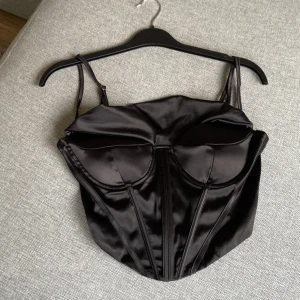 Svart satin korsett med snörning, M - Snygg svart korsett i satin med markerade kupor och smala axelband. Toppen har dekorativa sömmar framtill och dragkedja i ryggen för enkel på- och avtagning. Perfekt för att skapa en edgy och trendig look.oanvänd tror jag.storlek M.kort.vet inte märke