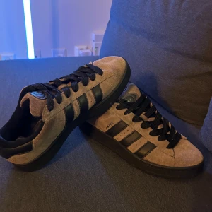 Adidas Campus sneakers (nypris 1599kr) i beige mocka storlek 44 - Säljer ett par Adidas Campus sneakers i beige mocka med svarta detaljer och klassiska tre ränder på sidan. Skorna har svarta snören, platt sula och rund tå. Perfekt för dig som gillar retrostil och vill ha något som sticker ut lite extra.