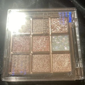 Glittrig ögonskuggspalett från Dikalu -Ny - Säljer en ögonskuggspalett med 9 skimrande och glittriga nyanser i brunt, rosa, silver och en holografisk glitterfärg. Paletten har fyrkantiga pans och kommer i en transparent plastförpackning. Perfekt för dig som älskar att experimentera med glansiga looks.