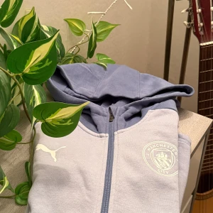 Manchester City hoodie från Puma ljusblå - Ljusblå hoodie från Puma med Manchester City-logga på bröstet och vit Puma-logga. Tröjan har dragkedja framtill och huva. Tillverkad i mjuk bomullsmix, perfekt för fotbollsfans som vill ha en chill och sportig stil. Litet fläck på men syns inte mycket för alla. Köpts från Zalando för 1000kr