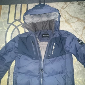 Blå Sail Racing dunjacka med huva - Säljer en mörkblå dunjacka från Sail Racing med huva och snygga detaljer. Jackan har dragkedja framtill, två bröstfickor med dragkedja och patch på ärmen med 'Sail Antarctica Expedition'. Insidan är ljusgrå och materialet känns slitstarkt och varmt.   Pris kan diskuteras vid snabb köp