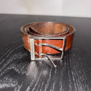 Brunt skärp från Tiger of Sweden - Snyggt brunt bälte från Tiger of Sweden i äkta skinn med en robust, fyrkantig metallspänne där märket är ingraverat. Perfekt accessoar för att lyfta din outfit med en klassisk och stilren touch.