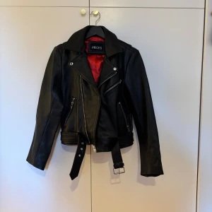 Svart skinnjacka från Pieces M - Snygg svart skinnjacka från Pieces i bikerstil med röd insida. Jackan har silvriga dragkedjor, bälte i midjan och klassisk krage. Perfekt för dig som vill ha en edgy look. Flera fickor med dragkedja och detaljer vid ärmslut. Nypris ca1500kr.              OBS! Endast hämtning. Bor i Sollentuna och Åkersberga 