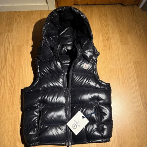 Svart dunväst från Moncler med huva - Snygg svart dunväst från Moncler med glansig finish, vadderad design och avtagbar huva. Västen har två fickor med dragkedja och klassisk Moncler-logga på bröstet. Perfekt för lager-på-lager och streetstyle.