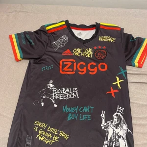 Ajax x Bob Marley fotbollströja Adidas - Unik svart Ajax fotbollströja från Adidas med Bob Marley-tema. Färgglada detaljer i rött, gult och grönt på ärmarna, tryck med Bob Marley, citat och graffitiliknande illustrationer. Ziggo-logga på bröstet och Ajax-märke. Tillverkad i lätt och ventilerande material.