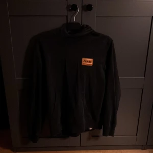 Svart Dickies hoodie med tryck - Svart hoodie från Dickies i storlek small med stor orange logga och coolt tryck på ryggen. Klassisk huva och känguruficka framtill. Skön och stilren, perfekt för dig som gillar streetwear och vill sticka ut lite extra.