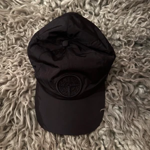 Svart keps från Stone Island - Säljer en svart keps från Stone Island med broderad logotyp framtill. Kepsen har en klassisk böjd skärm och justerbar snörning baktill. Tillverkad i lätt syntetmaterial som ger en sportig och clean look. Perfekt accessoar för dig som gillar streetwear.
