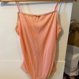 Rosa topp med spets och rosett - Supersöt ljusrosa body med tunna axelband, spetskant och liten rosett framtill. Toppen har ett diskret hålmönster i tyget och är perfekt för varma dagar. Materialet känns mjukt och luftigt, och toppen har en feminin och romantisk vibe.