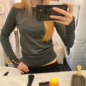 Mörkgrå fin stickad tröja Holly & Whyte - Mörkgrå stickad tröja från Holly & Whyte by Lindex i storlek XS. Tröjan har rund halsringning, långa ärmar och snygga dekorativa knappar i guld vid ärmsluten. 