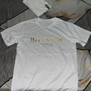 Vit Balmain Paris t-shirt - Snygg vit t-shirt från Balmain Paris med guldig logga framtill. Klassisk rund halsringning och korta ärmar. Tillverkad i mjuk bomull för en skön känsla. Perfekt för dig som gillar stilrena och exklusiva plagg.