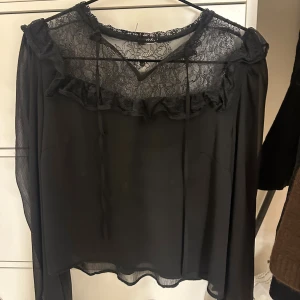 Svart blus med spets och volang - Säljer en svart blus med transparent tyg och vacker spets upptill. Blusen har volangdetaljer längs bröstet och knytband vid halsen. Långa ärmar i tunt material ger en luftig känsla. Perfekt för dig som gillar romantiska detaljer och en stilren look.