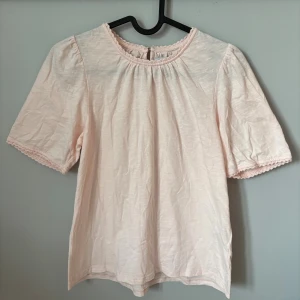 Ljusrosa blus från Holly & Whyte - Supersöt ljusrosa blus från Holly & Whyte med rund hals och korta puffärmar. Blusen har fina virkade detaljer vid halsringning och ärmslut. Tillverkad i mjuk bomull, perfekt för vår och sommar. Enkel och feminin stil som är lätt att matcha.