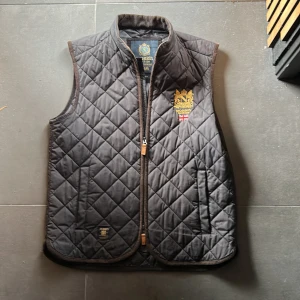 Svart quiltad väst från Morris - Quiltad svart väst från Morris med guldbroderad logga och brittisk flagga på bröstet. Västen har ståkrage, dragkedja framtill och två sidofickor. Materialet är polyester och designen har ett klassiskt rutmönster. Perfekt för dig som gillar stilren och tidlös look.
