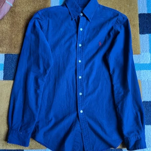 Blå skjorta från Ralph Lauren - Klassisk blå skjorta från Ralph Lauren med lång ärm och knappar framtill. Skjortan har en röd broderad logga på bröstet och är tillverkad i mjuk bomull. Perfekt för dig som gillar stilrena och tidlösa plagg.