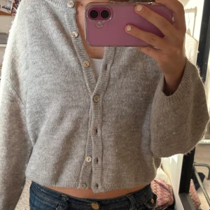 Grå kofta från Gina Tricot - Mysig grå kofta från Gina Tricot med rund halsringning och knappar framtill. Koftan är stickad och har en lite croppad passform med långa ärmar. Perfekt att slänga på sig över ett linne eller t-shirt.