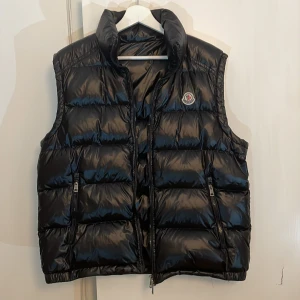 Moncler väst - Snygg och klassisk Moncler-väst i blank svart färg. Vadderad med dun som håller värmen utan att kännas tung. Stilren modell med dragkedja, två fickor med zip samt ikoniskt Moncler-märke på bröstet. Priset kan diskuteras.