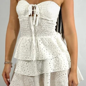 Vit klänning med volanger - Supersöt vit kortklänning med broderade hålmönster, volanger och omlottknytning framtill. Axelbandslös modell med smockad midja och flera lager i kjolen för extra volym. Perfekt för sommarens alla tillfällen!