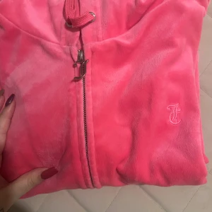 Juicy Couture rosa velour hoodie XL - Säljer en ikonisk rosa hoodie från Juicy Couture i mjuk velour. Tröjan har dragkedja med snygg JC-detalj, huva med snörning och en broderad logga på bröstet. Perfekt för dig som vill ha den där klassiska Y2K-looken.
