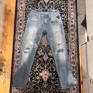 Replay Anbass Jeans 28/32, blå tvätt med slitningar - SäljerEtt par Replay Anbass jeans i storlek 28/32 letar nu efter en ny ägare. Dessa jeans har en snygg blå tvätt med trendiga slitningar som ger den där avslappnade, moderna looken utan att kännas för slitna.  Modellen Anbass är en av Replay’s mest populära, just för att den kombinerar bekvämlighet med en smickrande passform. Slim fit som sitter bra längs benen, normal midja och en stil som funkar i alla sammanhang. Ett riktigt mångsidigt par jeans som kan bäras året runt.