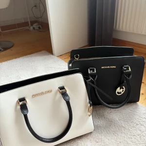 Vit och svart handväska från Michael Kors - Snygga och stilrena handväskor från Michael Kors i vit och svart med guldfärgade detaljer. Väskan har två svarta handtag, innerfack med dragkedja och en klassisk rektangulär form. Perfekt för dig som gillar minimalistisk design och vill ha plats för allt viktigt. Helt oanvänd då jag köpt fel modell. Går att köpa enskilt eller båda. Skickas mot frakt kostnad. 