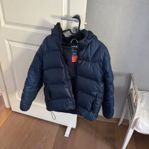 Mörkblå pufferjacka från Helly Hansen - Säljer en mörkblå pufferjacka från Helly Hansen med huva och dragkedja. Jackan har två sidofickor med dragkedja och reflekterande detalj på ärmen. Perfekt för kalla dagar och riktigt skön att bära. Har även två små hål men är i bra skick.