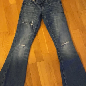 Blå bootcut jeans med slitningar - Säljer ett par blå jeans med bootcut passform low waist och slitna detaljer på framsidan av benen. Jeansen har klassiska fickor både fram och bak, och är tillverkade i jeansmaterial. Perfekt för dig som gillar en avslappnad och trendig stil.