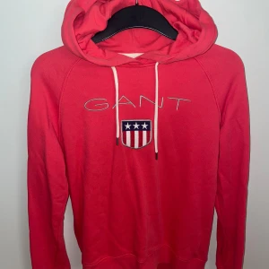 GANT rosa/röd hoodie - Säljer en snygg rosa/röd hoodie från GANT med broderad logga och sköld på bröstet. Tröjan har huva med vita snören och långa ärmar. Perfekt för dig som gillar klassisk och sportig stil. Är i ett fint skick, och säljer då den är för liten för mig men oxå kommer inte till användning. Vid frågor hör av er💕