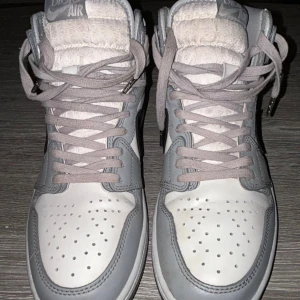 Nike Air Jordan 1 Dior Highs - Äkta, exklusiva Nike Air Jordan 1 Dior Highs i vit och ljusgrå skinn med Dior-mönstrad swoosh och genomskinlig sula med Dior-logga. Skorna har snörning, perforerad tå och högt skaft för en riktigt lyxig vibe. Perfekt för dig som vill sticka ut med limited edition sneakers. Pris kan diskuteras!